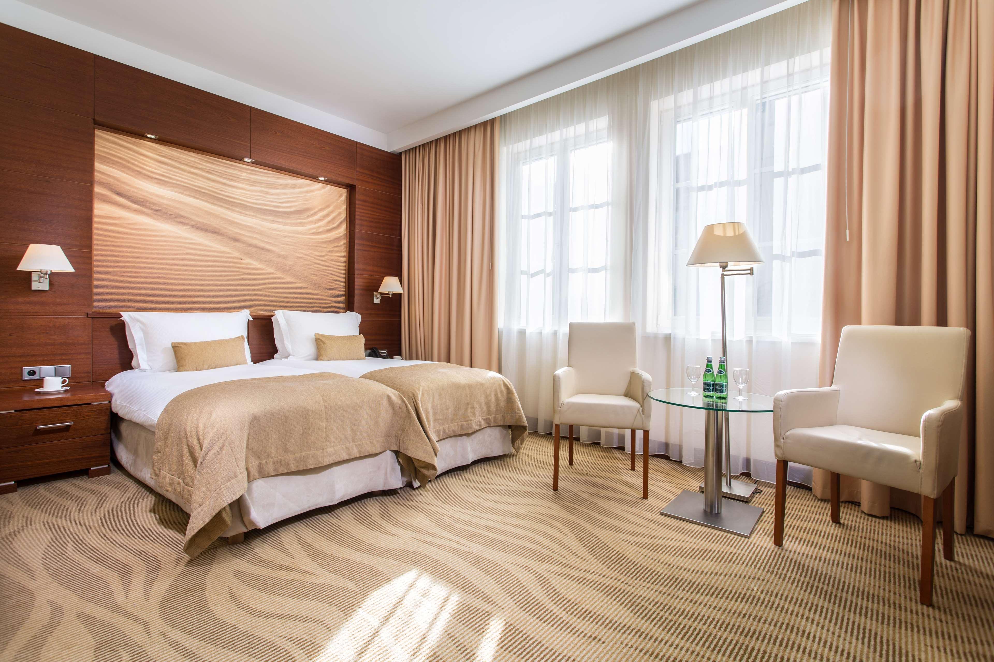 Radisson Blu Hotel, Danzica Esterno foto The photo shows a well-decorated hotel room featuring two twin beds with beige linens and pillows. The walls have a warm, wooden paneling accent, and large windows are dressed with light curtains allowing natural light to fill the space. There are tw
