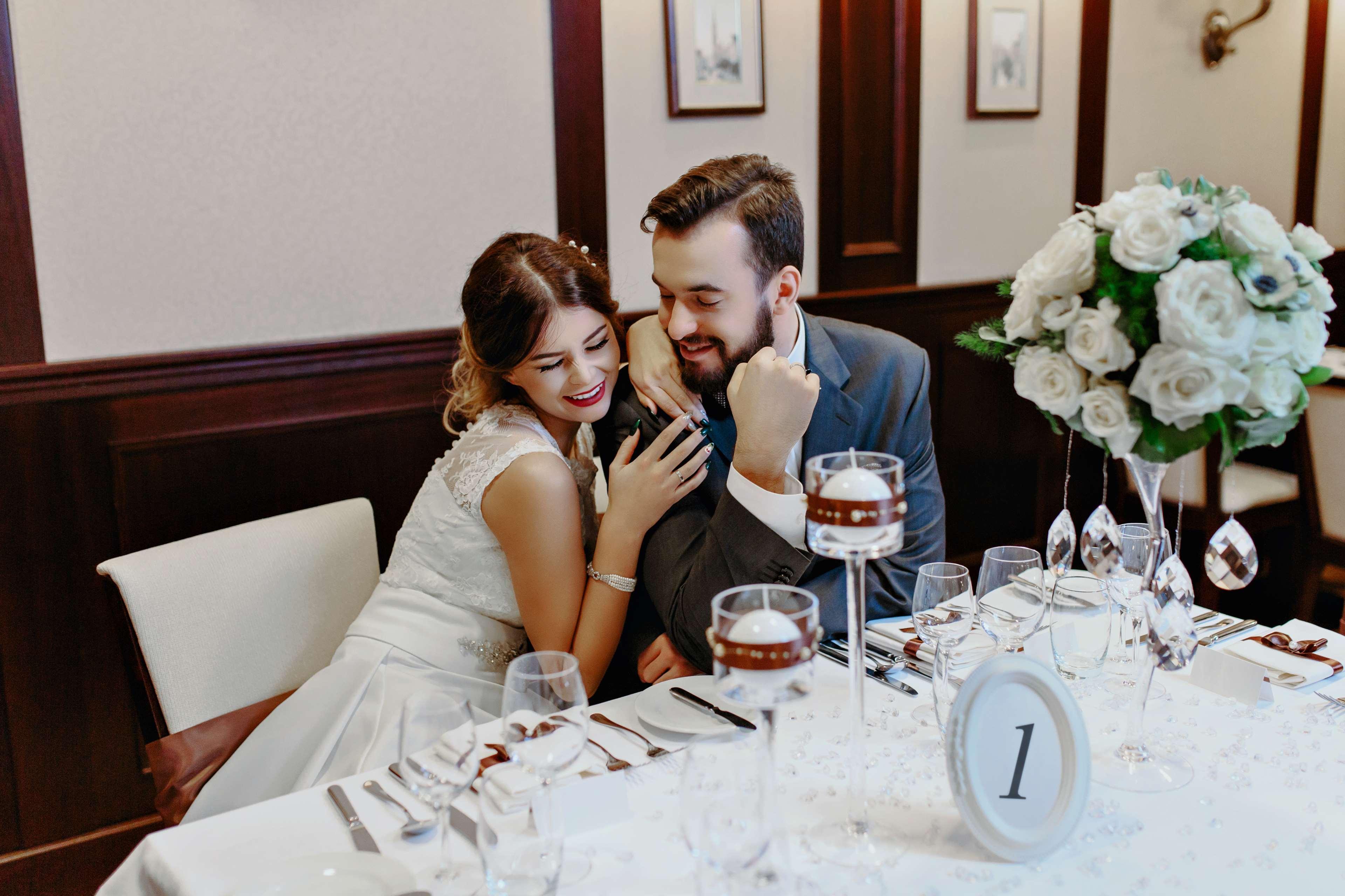 Radisson Blu Hotel, Danzica Esterno foto The photo depicts a couple sitting at a beautifully arranged dining table. The woman, dressed in a white wedding gown, leans affectionately towards the man, who is wearing a gray suit. They appear to be enjoying a moment together, smiling and engagin