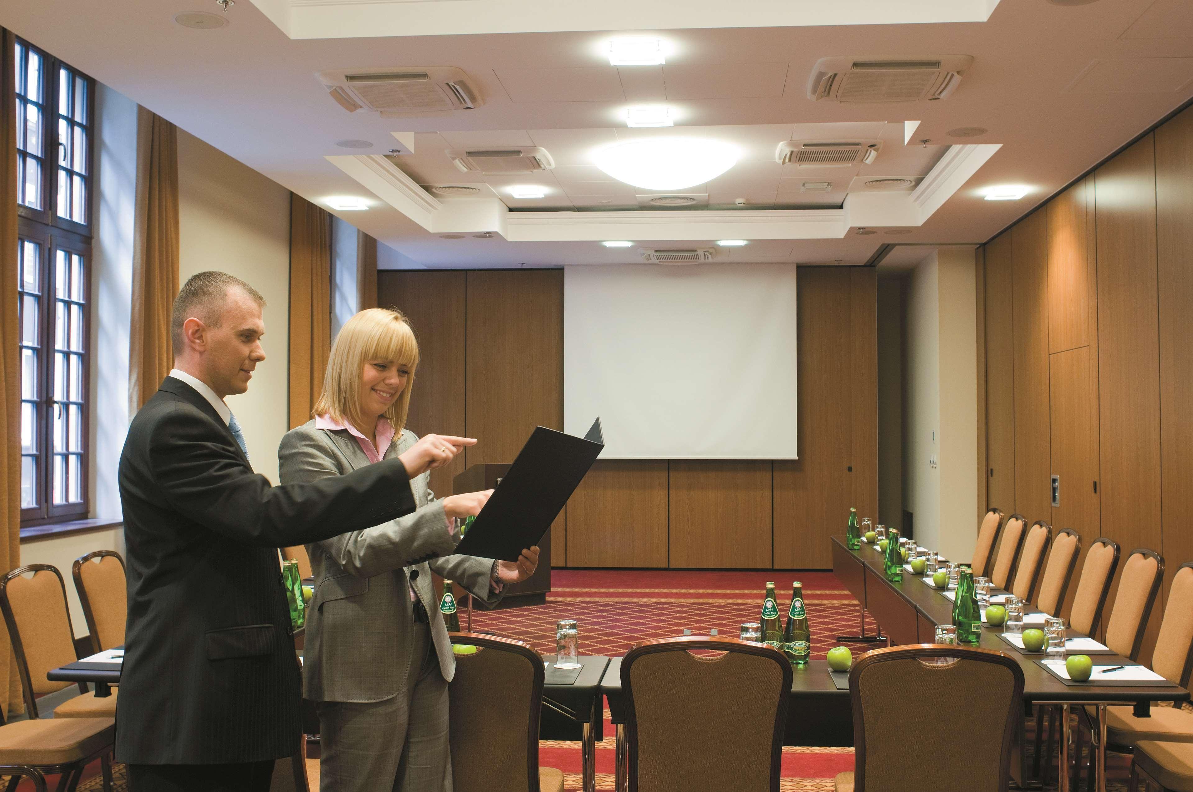 Radisson Blu Hotel, Danzica Servizi foto The image shows a professional meeting room set up for a conference or presentation. There are two people in formal attire engaged in discussion; one is holding a document or folder while the other is pointing at it. The room features a large screen 