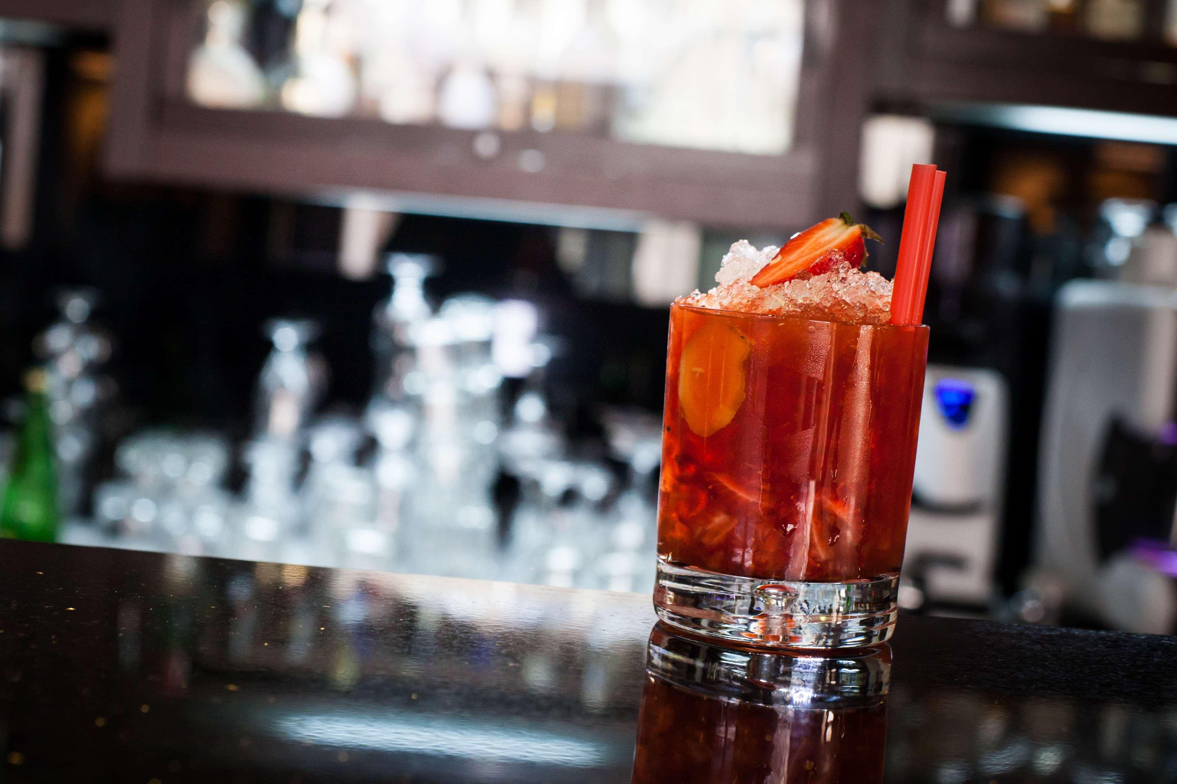 Radisson Blu Hotel, Danzica Esterno foto The photo shows a glass of a colorful cocktail placed on a dark surface, likely a bar countertop. The drink is garnished with slices of fruit, possibly strawberries, and is filled with ice. In the background, there are blurred outlines of other glass
