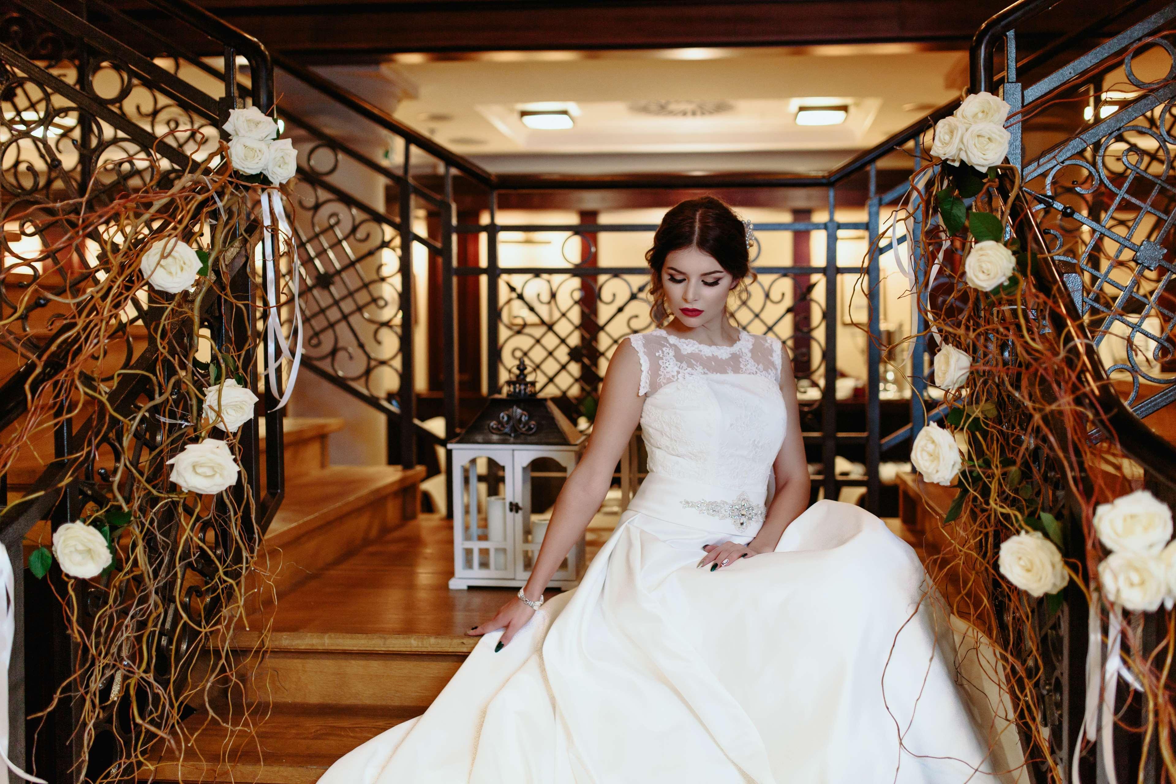 Radisson Blu Hotel, Danzica Esterno foto The image shows a bride seated on a staircase in an elegant indoor setting. She is wearing a beautiful white wedding gown with intricate details, including a sheer neckline. The staircase is adorned with decorative elements, such as white roses and f