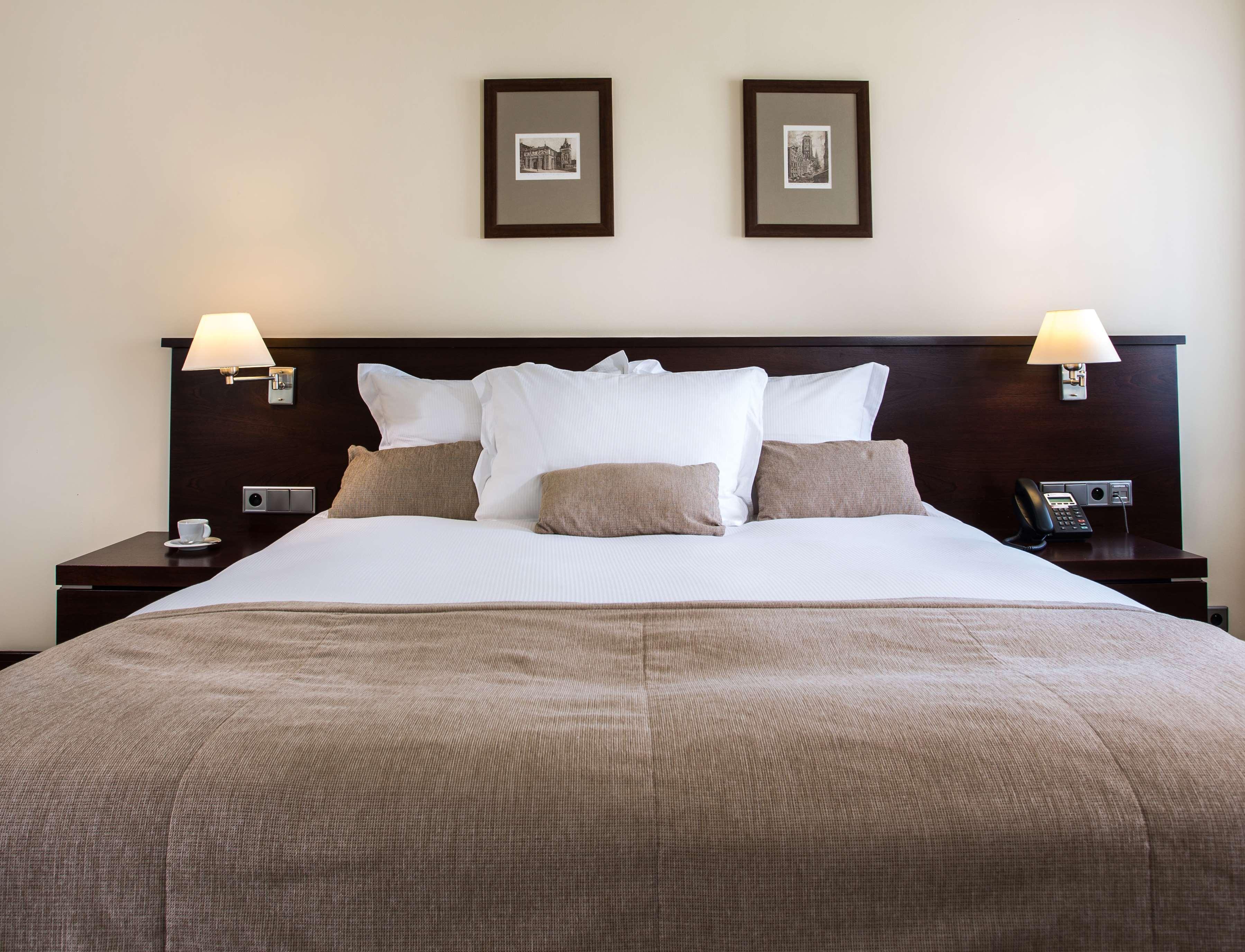 Radisson Blu Hotel, Danzica Esterno foto The photo shows a well-decorated hotel room, focusing on a large bed with a dark wood headboard. The bed is adorned with several white pillows and a couple of beige cushions. There are two lamps on either side of the bed, providing soft lighting. Abo