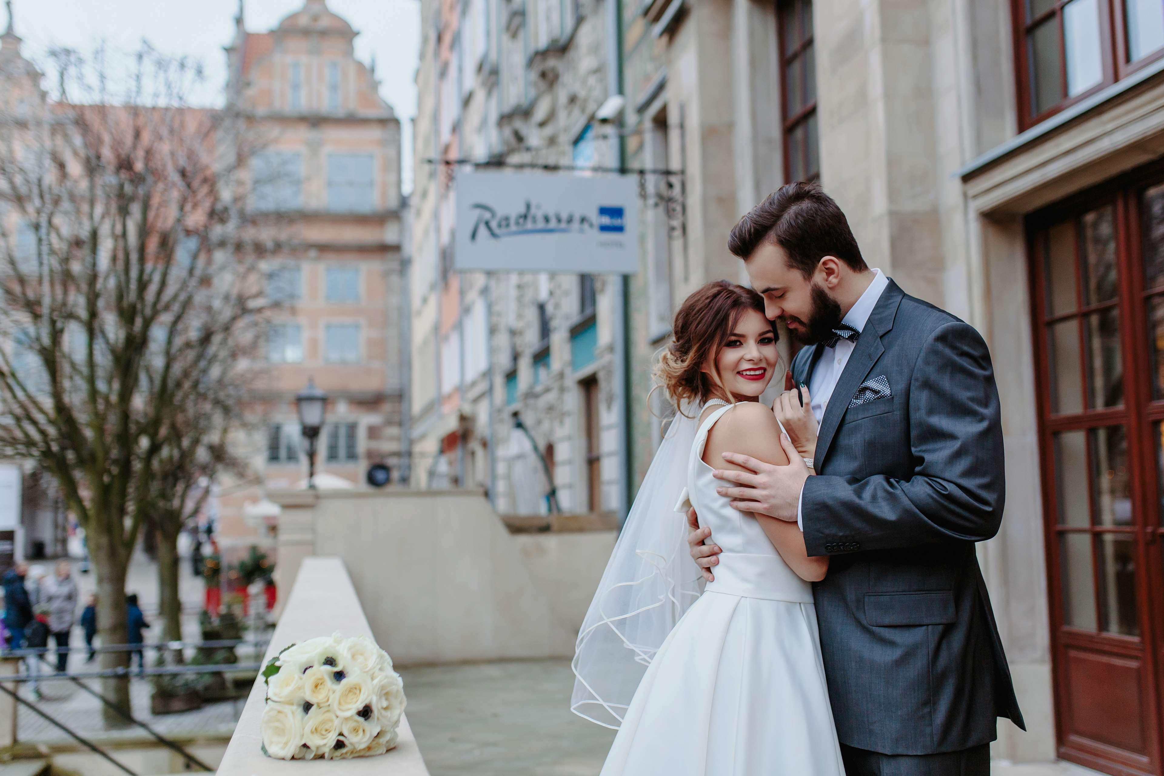 Radisson Blu Hotel, Danzica Esterno foto The photo depicts a bride and groom embracing on a balcony. The bride is wearing a white wedding dress with a veil, and she is smiling while leaning into the groom. He is dressed in a grey suit and is holding her close, both appearing very happy. In 