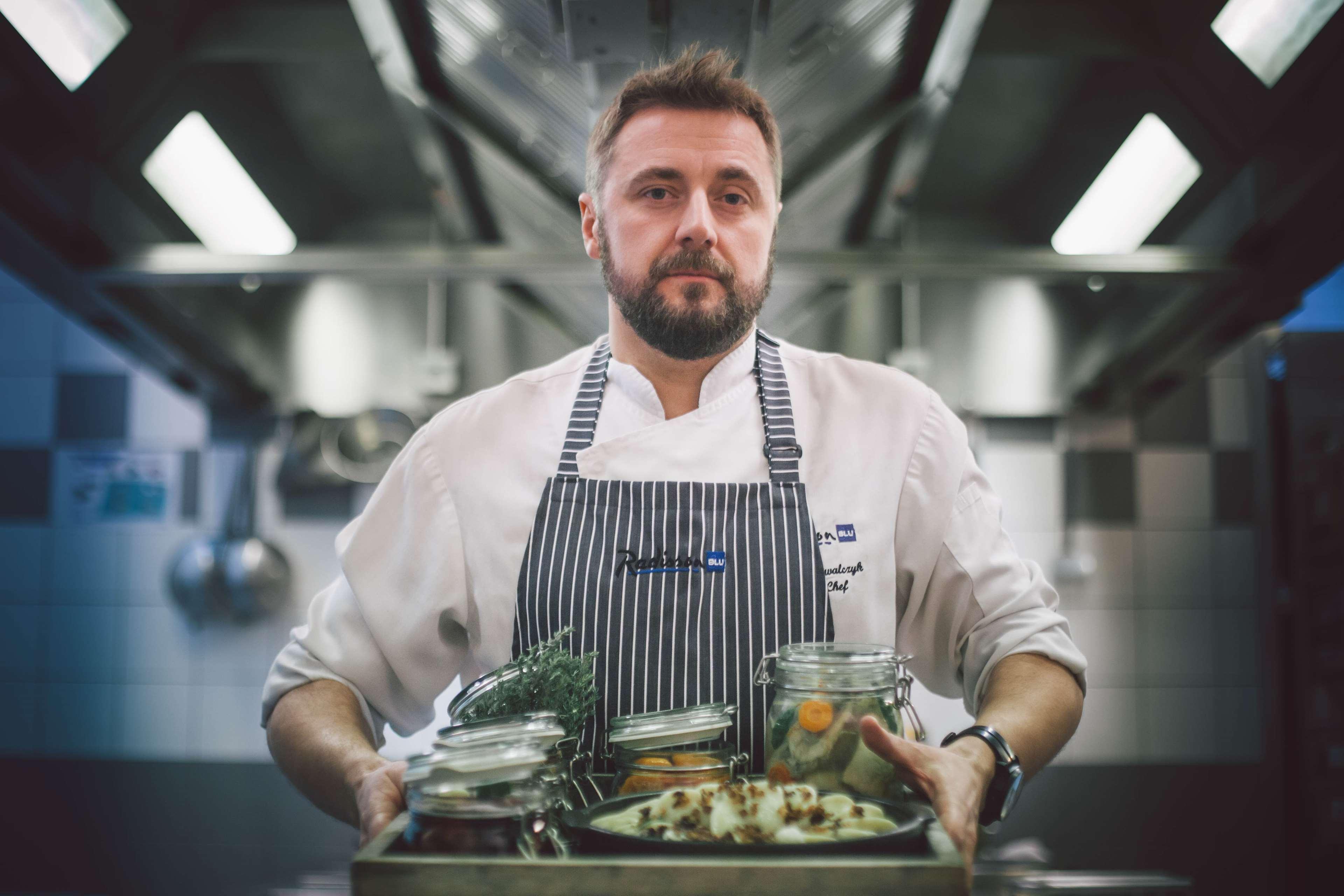 Radisson Blu Hotel, Danzica Esterno foto The photo depicts a chef in a professional kitchen setting. He is wearing a white chef's coat with a striped apron and has a focused expression. He is holding a wooden tray filled with various jars and containers that likely contain ingredients for c