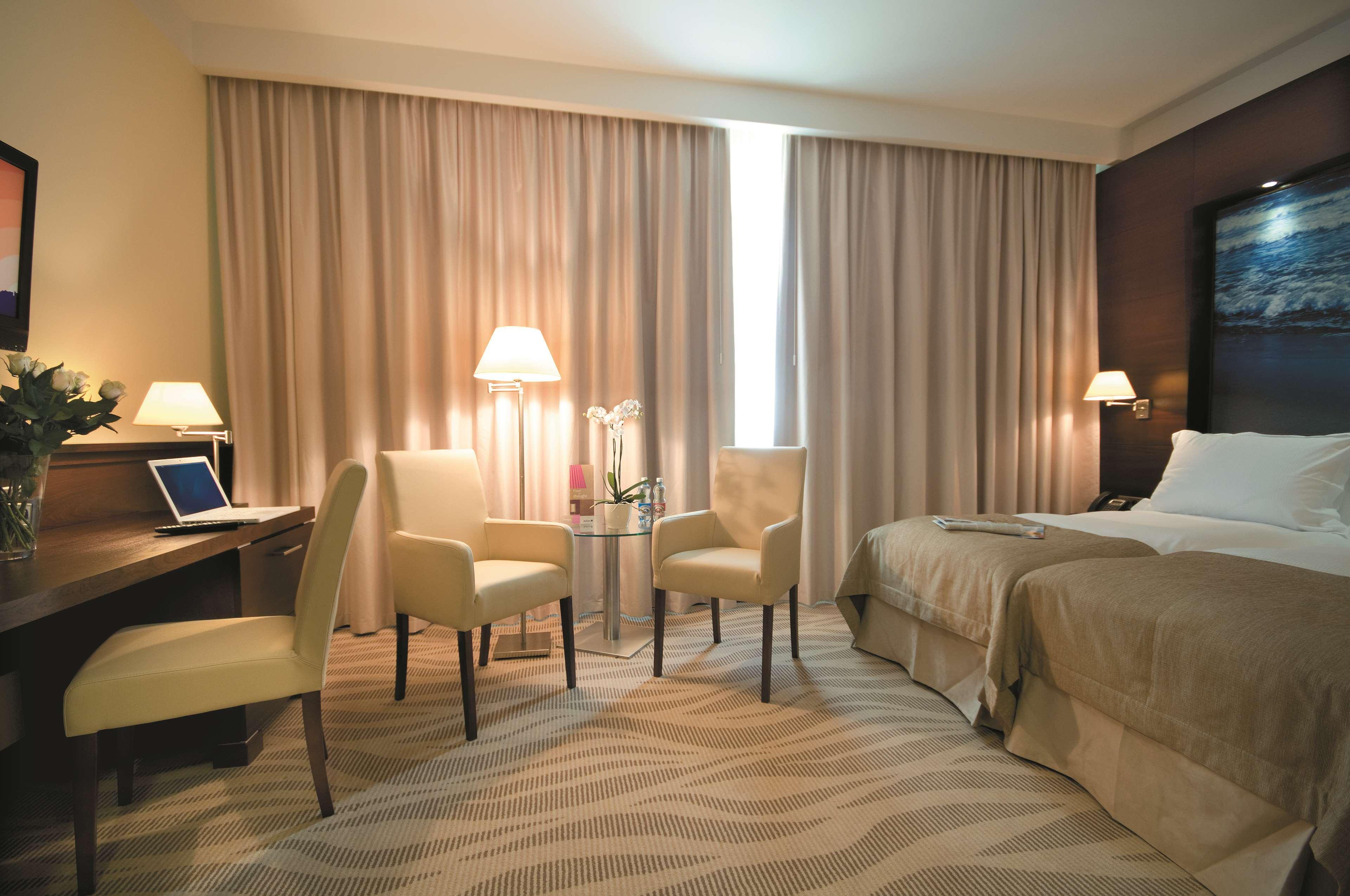 Radisson Blu Hotel, Danzica Esterno foto The photo shows a hotel room that features a queen-sized bed with neutral-colored bedding. There are two chairs and a small glass table in front of the bed. A desk is situated to one side, likely for work or personal use. The room has two lamps provi