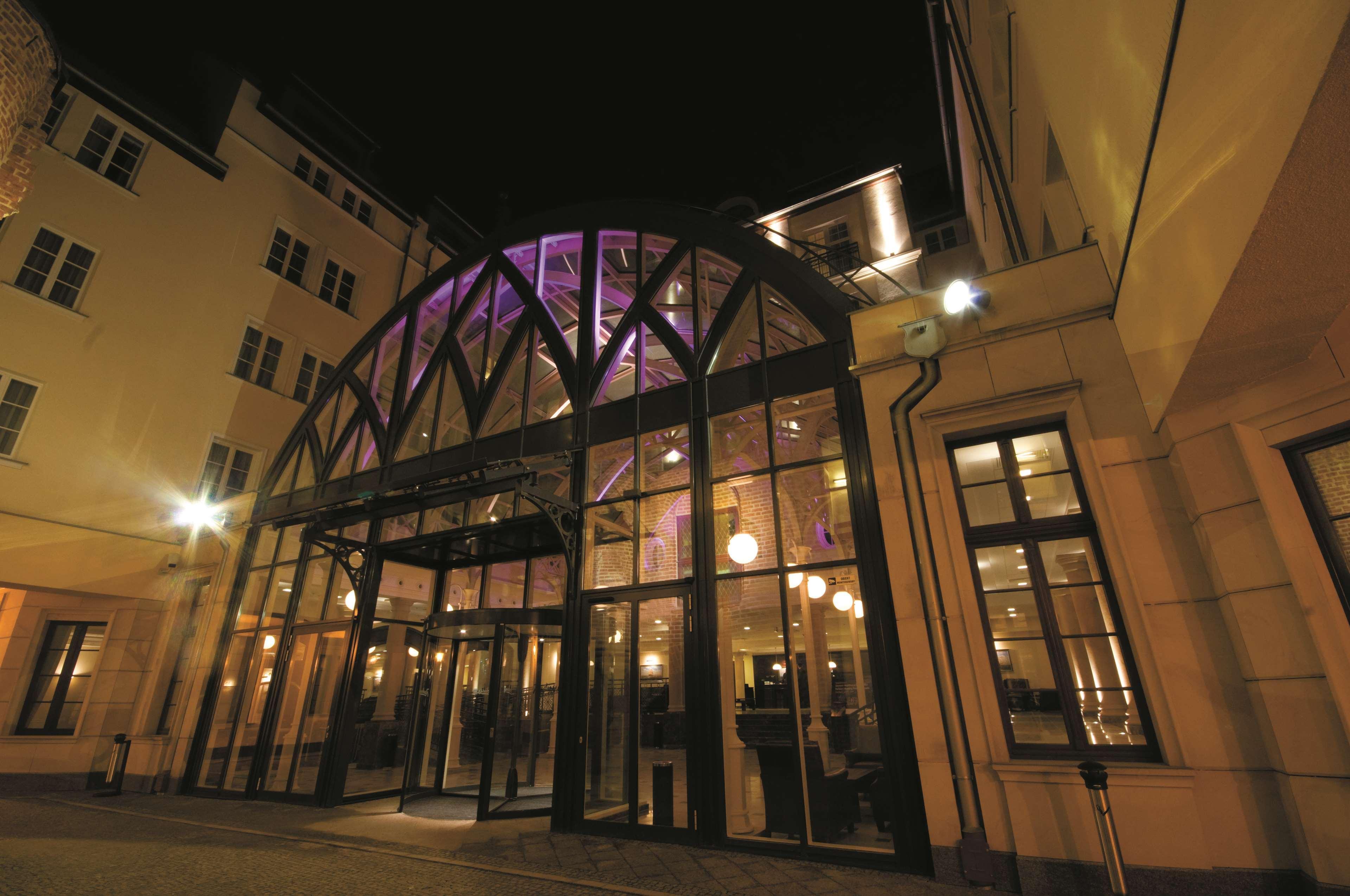 Radisson Blu Hotel, Danzica Esterno foto The photo shows the entrance of a building at night. The structure features a large glass facade with an arched roof supported by a metal framework. The lights illuminate the entrance and the interior, creating a welcoming atmosphere. Soft purple lig
