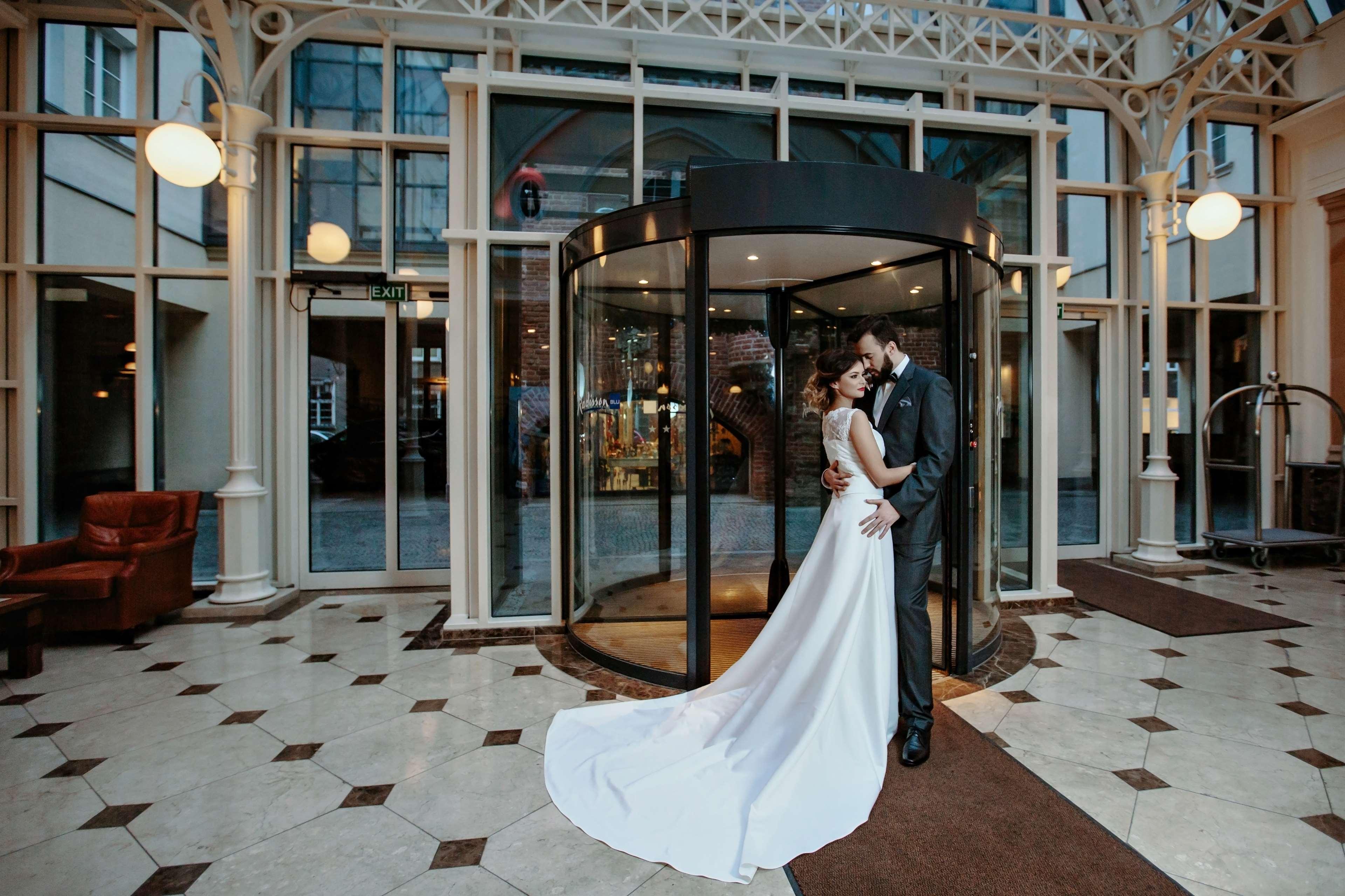 Radisson Blu Hotel, Danzica Esterno foto The photo shows a couple in formal attire standing outside a building, likely a hotel or venue. They appear to be newlyweds, with the woman in a beautiful white wedding gown and the man in a suit. They embrace each other tenderly in front of a glass 