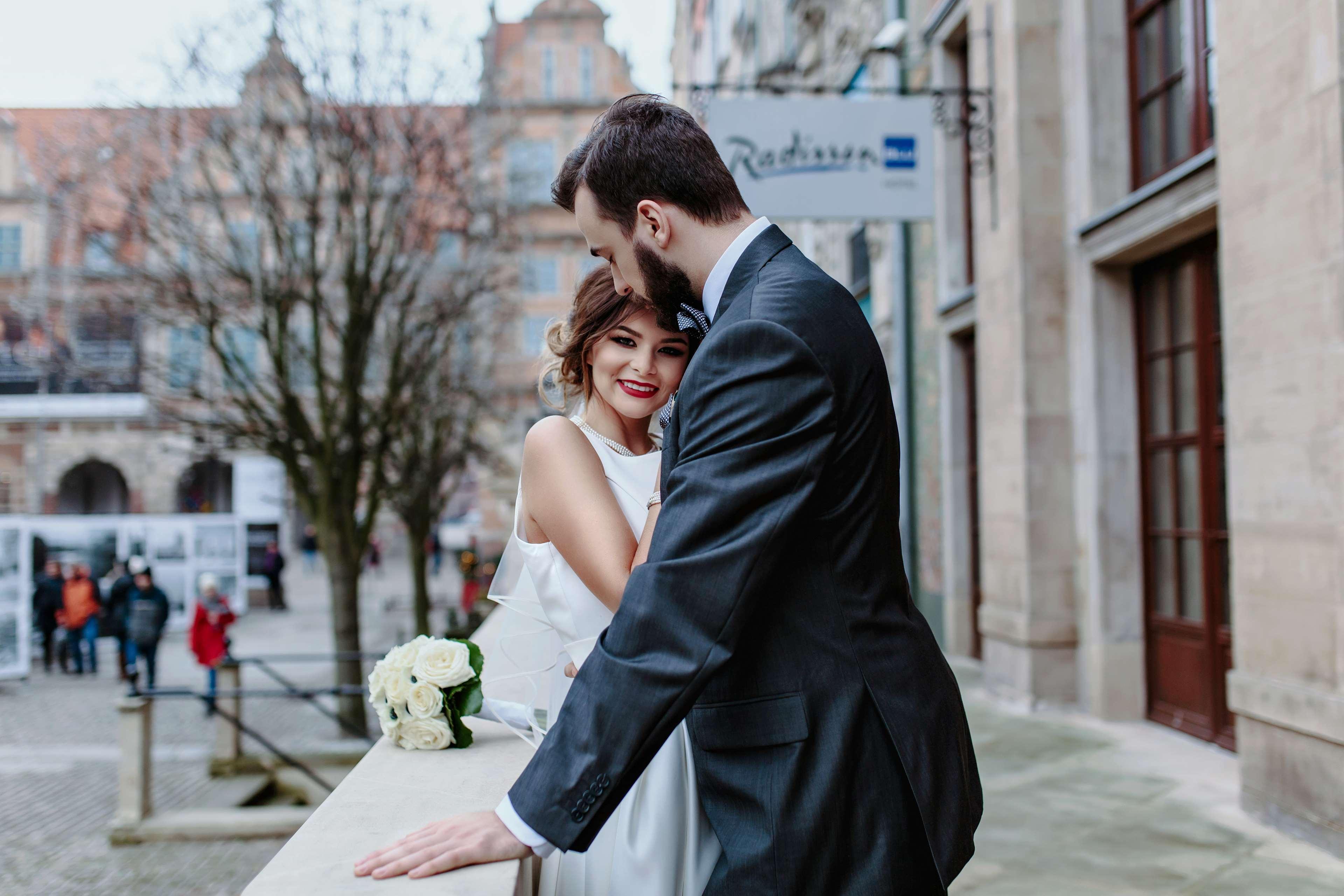 Radisson Blu Hotel, Danzica Esterno foto The photo shows a couple embracing in an outdoor setting. The woman, dressed in a white wedding gown, is smiling and appears to be enjoying the moment. The man, wearing a dark suit, leans in toward her, suggesting a romantic atmosphere. In the backgr