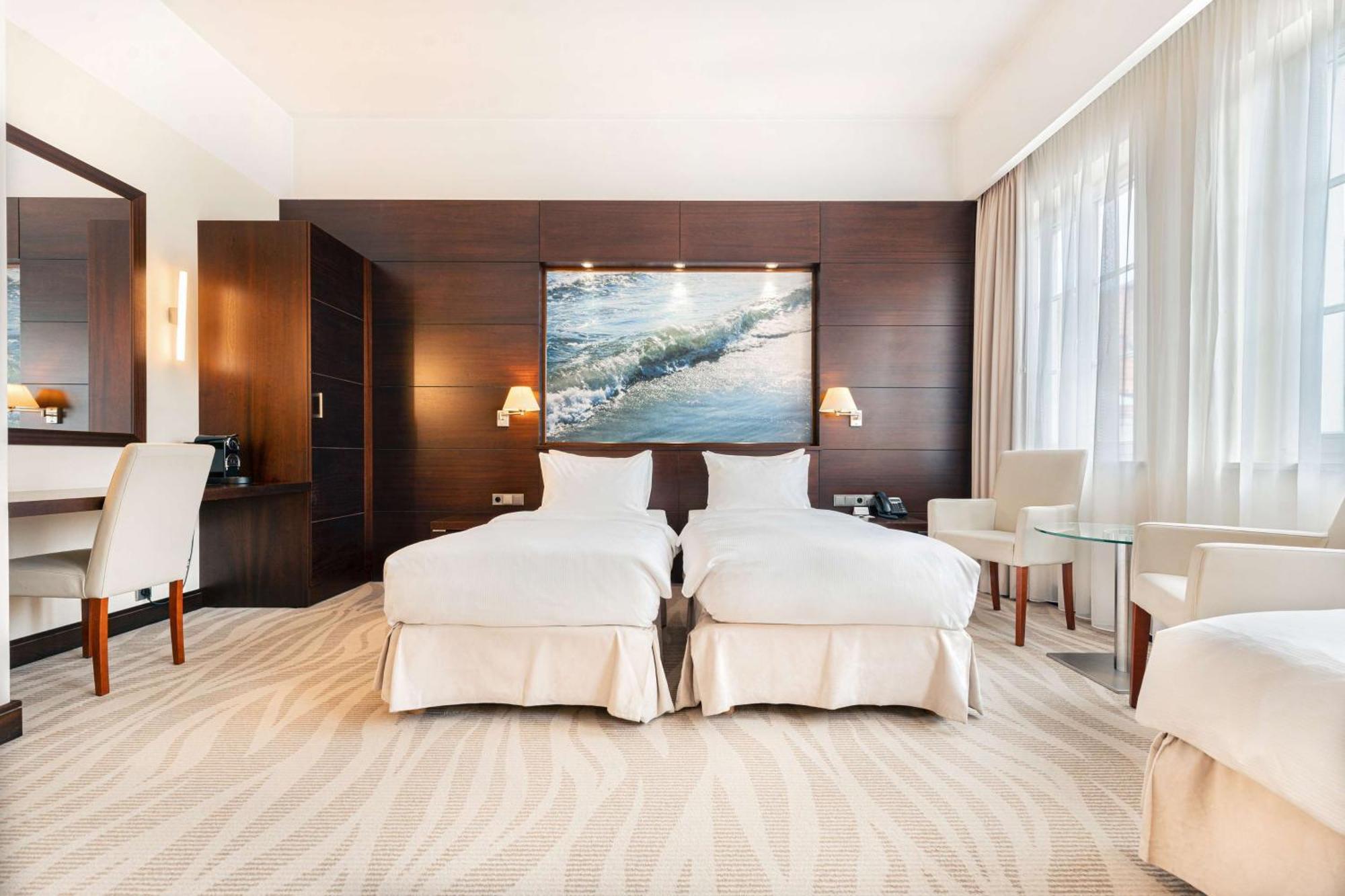 Radisson Blu Hotel, Danzica Esterno foto The photo shows a modern hotel room featuring two twin beds dressed in white bedding. The room has a warm, elegant design with wooden cabinetry on one wall and a large piece of artwork depicting a landscape or seascape above the beds. There are two b
