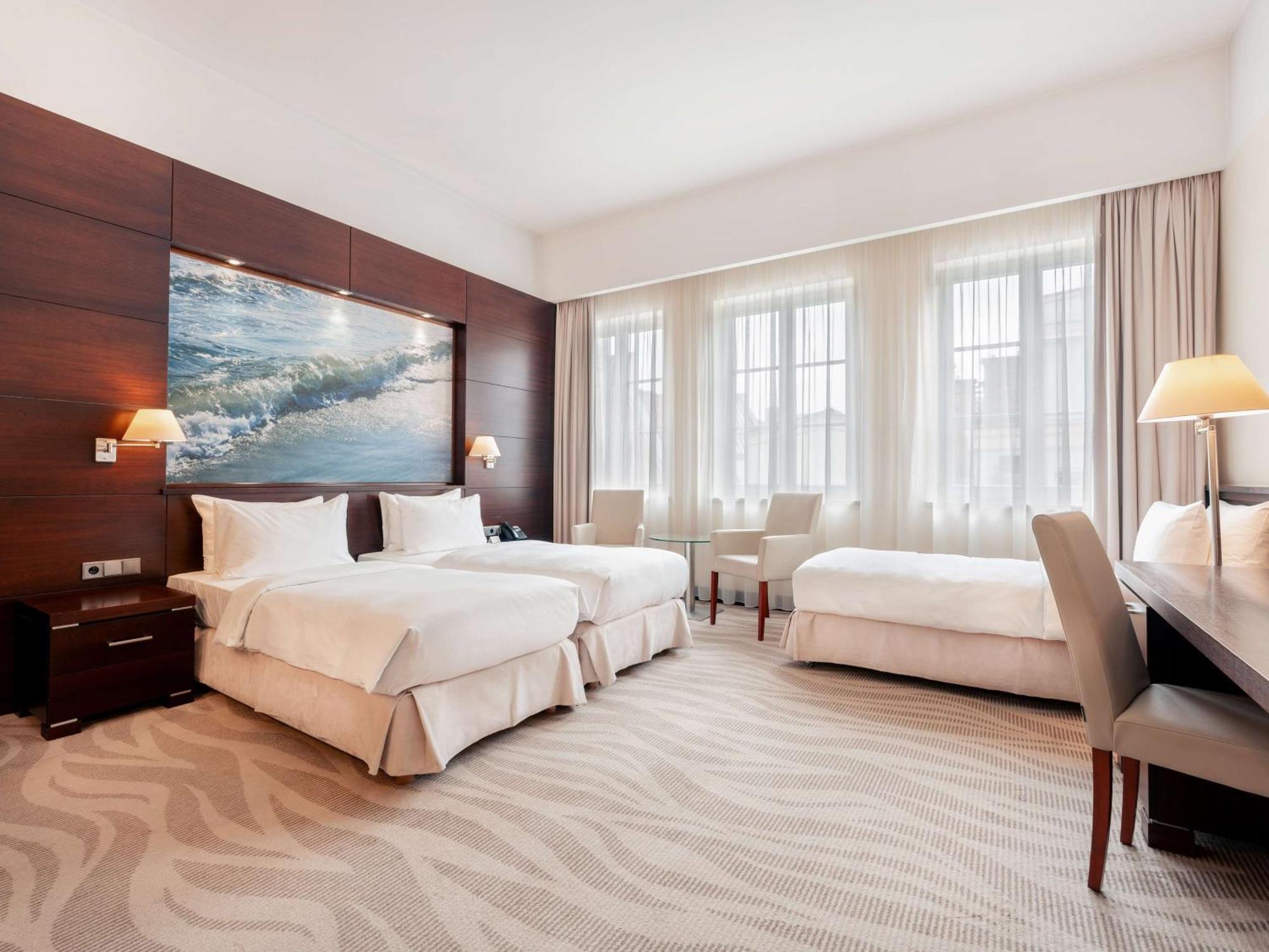 Radisson Blu Hotel, Danzica Esterno foto The photo depicts a modern hotel room featuring two twin beds with white bedding, arranged against a wooden wall that includes a large artwork of ocean waves. The room is brightened by natural light streaming through three large windows, which are ad