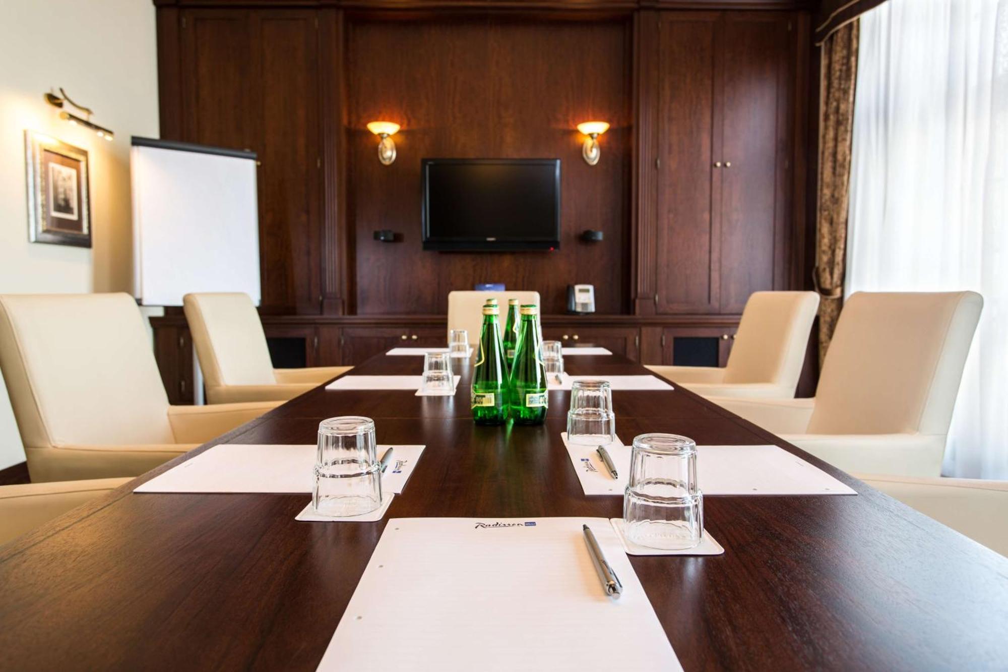 Radisson Blu Hotel, Danzica Esterno foto The photo shows a conference room setup. There's a large wooden table with several cream-colored chairs around it. On the table, there are sheets of paper, water glasses, and small bottles of water. A flat-screen television is mounted on the wall at 