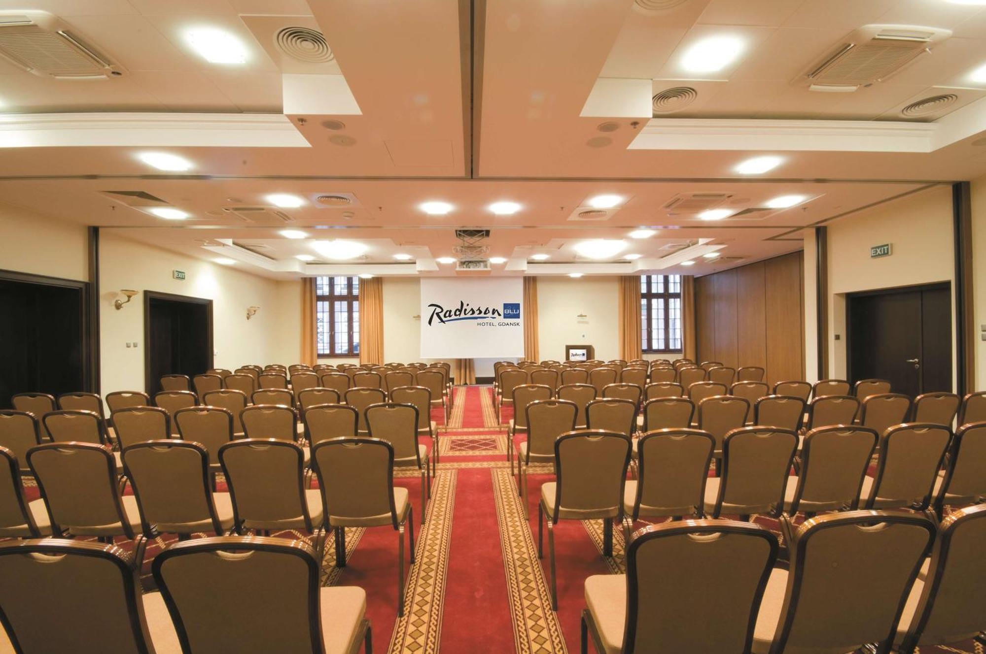 Radisson Blu Hotel, Danzica Esterno foto The photo shows the interior of a conference room or event space. It features rows of brown chairs arranged neatly, facing towards the front where a stage or presentation area is set up. There is a carpet running down the center of the room, and the 