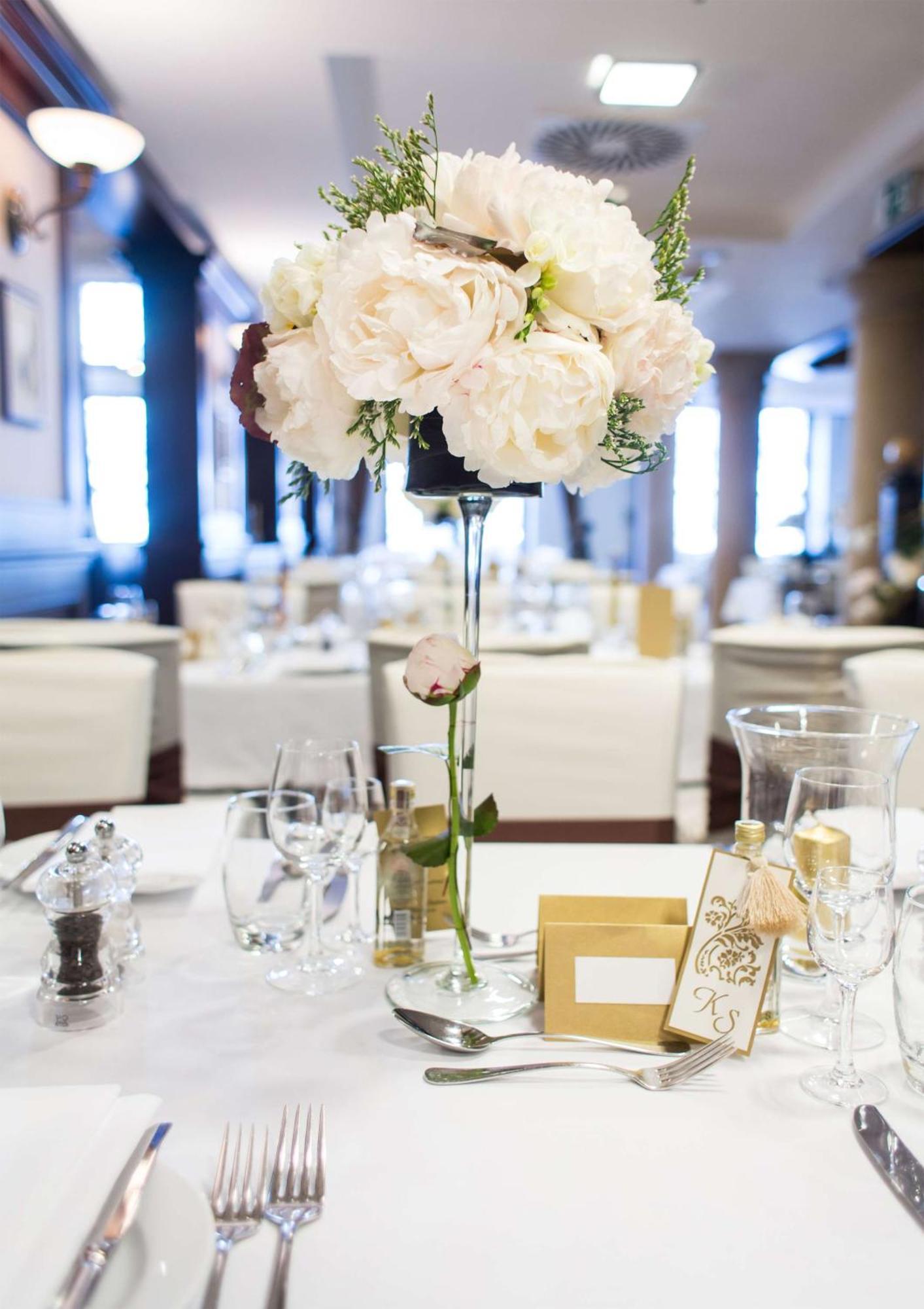 Radisson Blu Hotel, Danzica Esterno foto The photo depicts a beautifully arranged table setting, likely for a formal event or celebration. At the center of the table is a tall floral arrangement featuring soft pink peonies and greenery, elegantly placed in a clear vase. Surrounding the cent