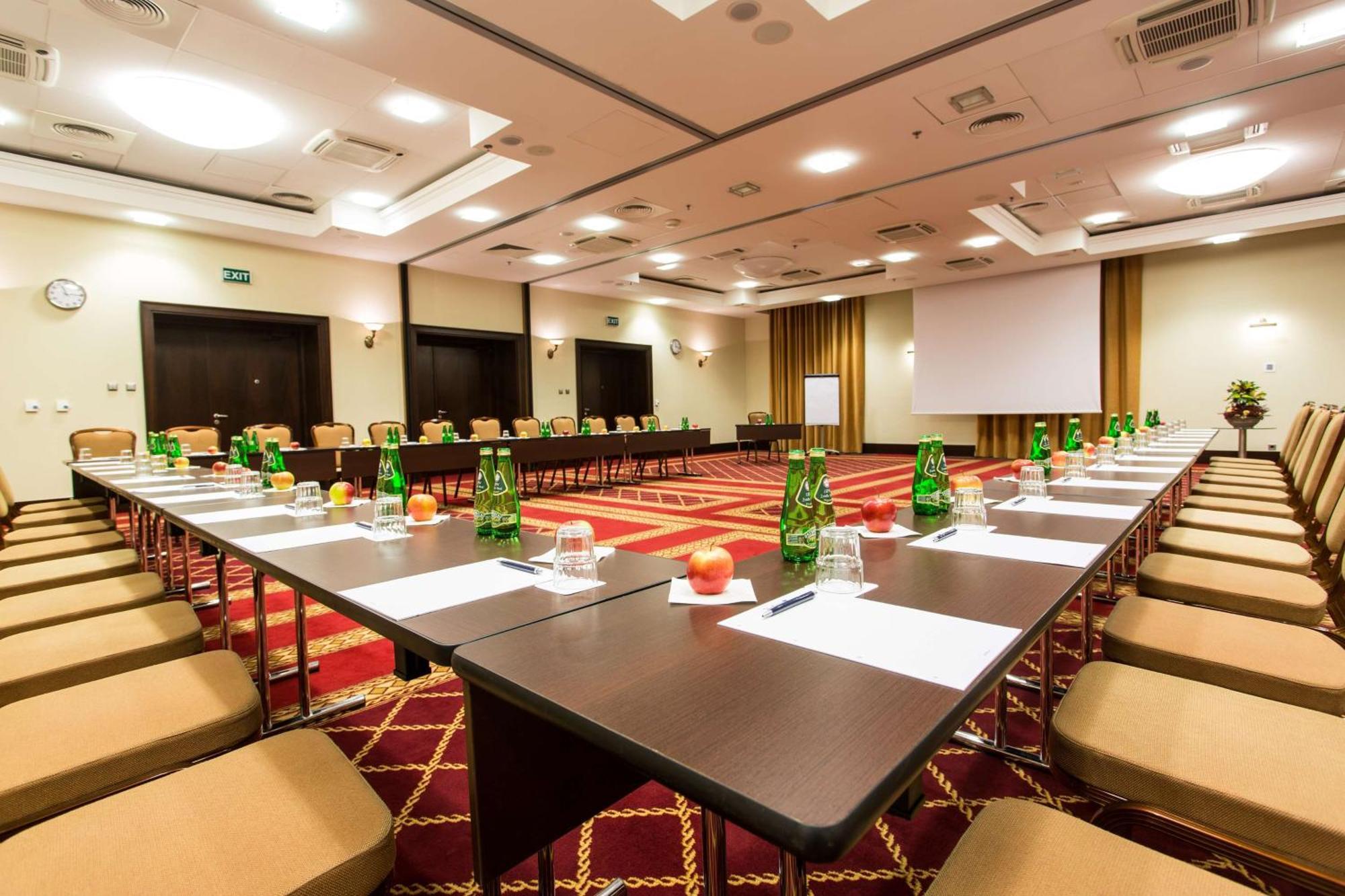 Radisson Blu Hotel, Danzica Esterno foto The photo shows a conference room set up for a meeting or seminar. Long tables are arranged in a U-shape, surrounded by beige chairs. Each table has green glass bottles of water, small glasses, and apples placed on it. There are also sheets of paper 