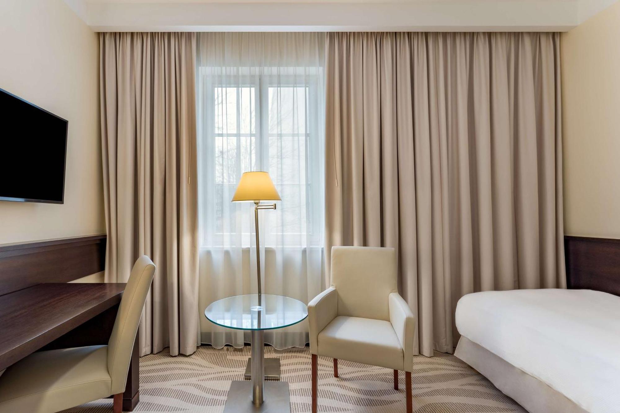 Radisson Blu Hotel, Danzica Esterno foto The photo shows a hotel room that features a light and neutral color scheme. There is a window covered with sheer curtains, allowing natural light to enter the space. To the right, there is a bed with a white bedspread. 

In the center, there is a sm