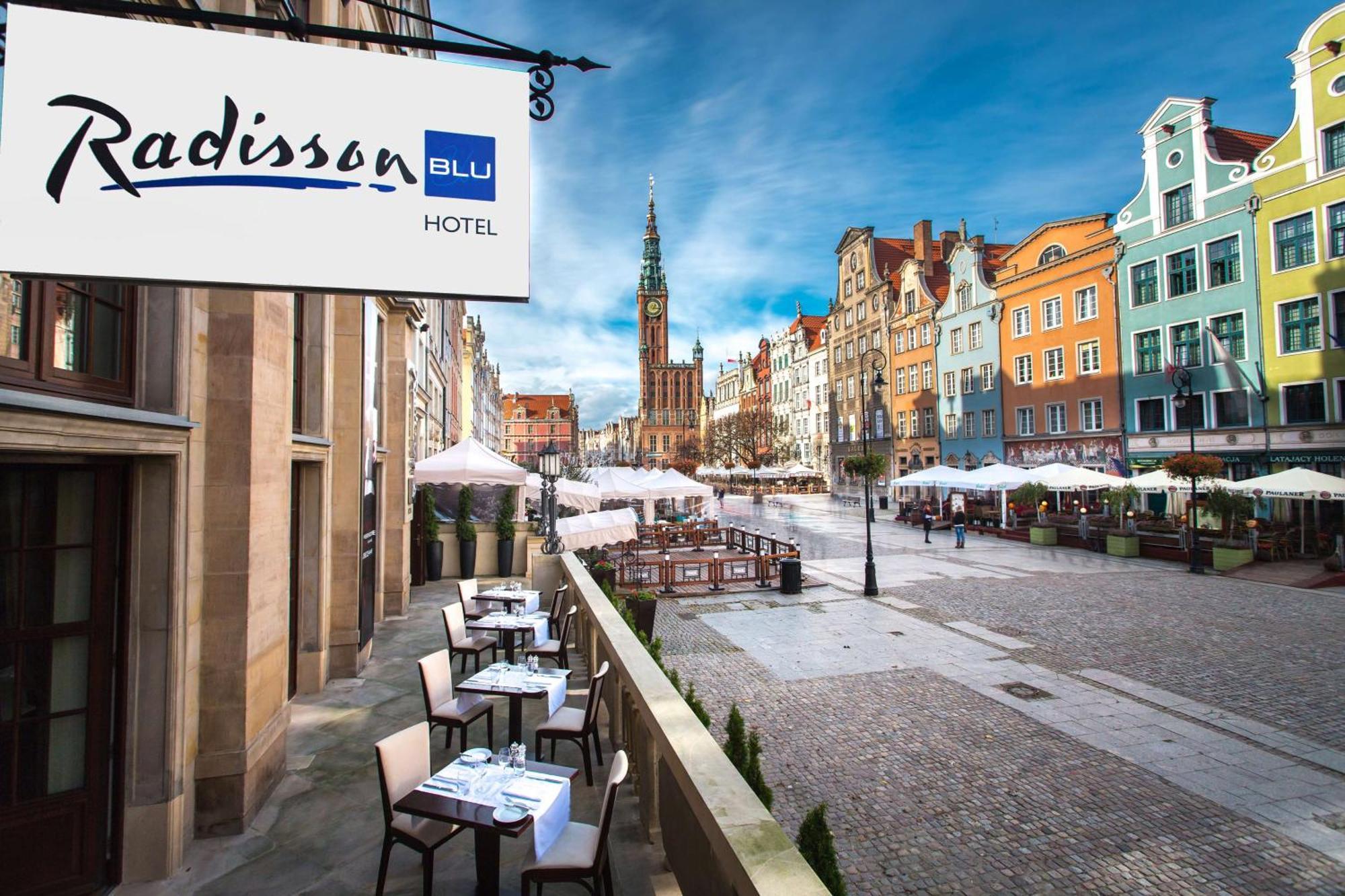 Radisson Blu Hotel, Danzica Esterno foto The photo shows a view of the Radisson Blu Hotel, which is situated in a vibrant, picturesque area. The image features an outdoor seating arrangement with tables and chairs, likely part of a restaurant or café. In the background, colorful historic bu