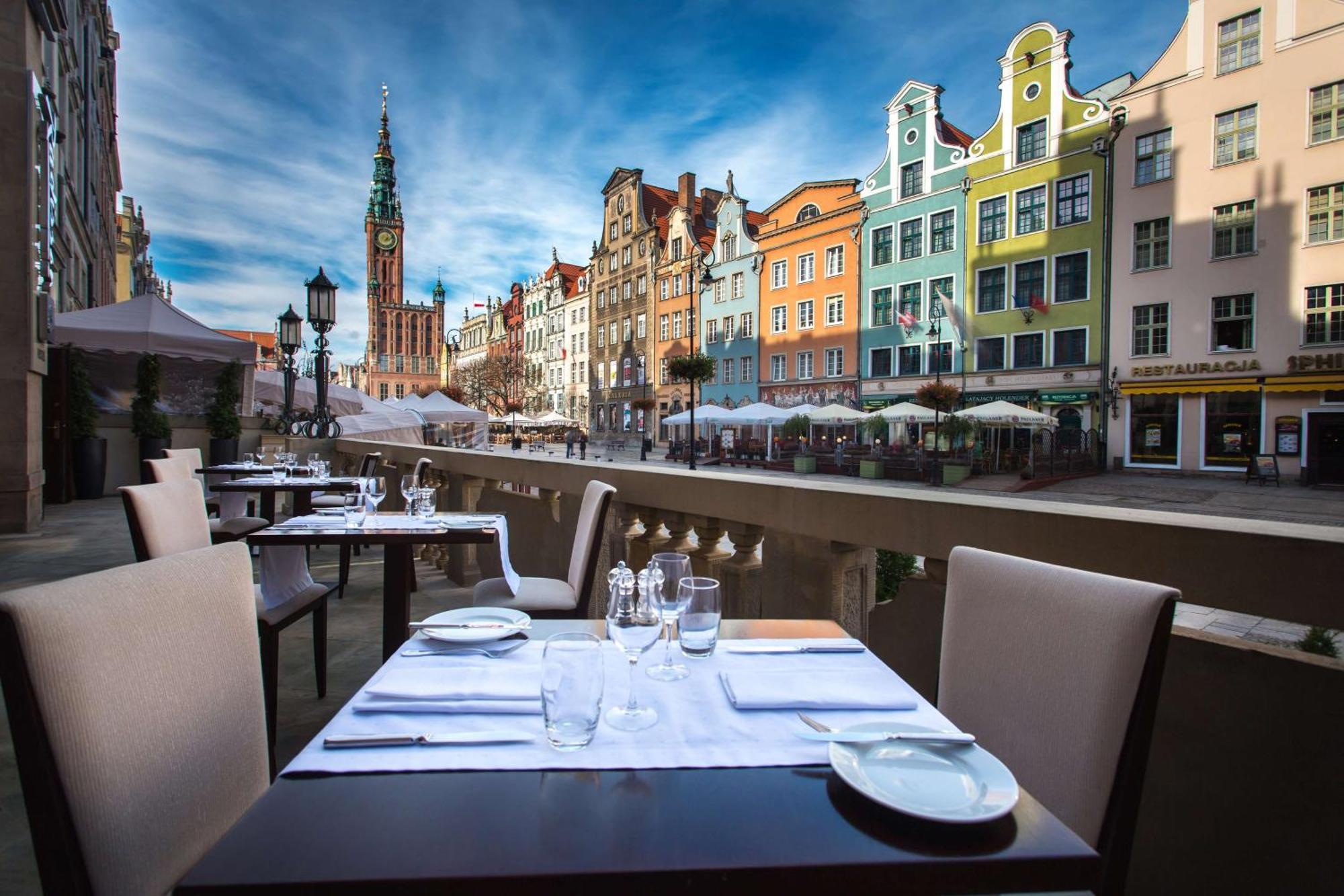 Radisson Blu Hotel, Danzica Esterno foto The photo shows an outdoor dining area with elegantly set tables overlooking a picturesque street lined with colorful buildings. In the background, there is a tall clock tower, suggesting a historic or charming town. The sky is bright with some cloud