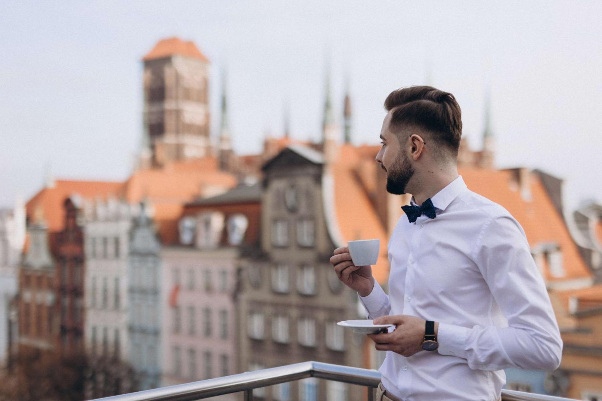 Radisson Blu Hotel, Danzica Esterno foto The photo features a man standing on a balcony, holding a cup of coffee or tea in one hand while resting a saucer in the other. He is dressed in a white shirt with a bow tie. The background reveals a picturesque view of an old town, showcasing buildi